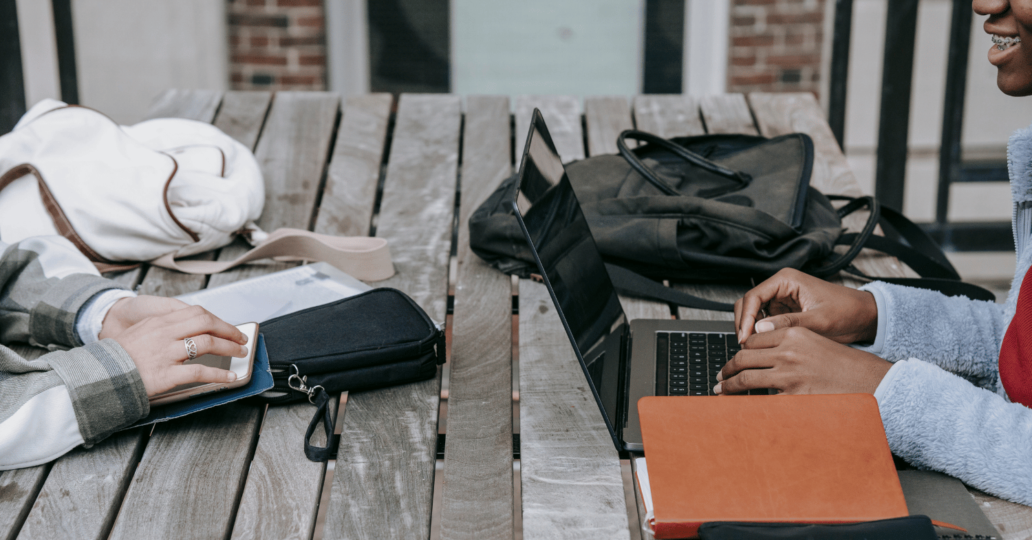 Two students sitting opposite on a wooden table, one working on a laptop and one working on a tablet